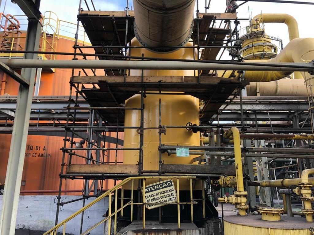 Coluna com andaime em planta de amônia, ao lado de tanque com placa de aviso de vazamento